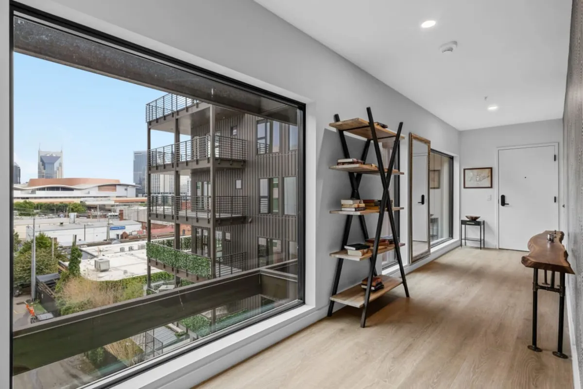 A bright interior scene with expansive windows revealing a city view, a bookshelf filled with books, and a welcoming entryway. The abundant natural light and urban outlook evoke a sense of downtown living and connectivity to Nashville's vibrant atmosphere.