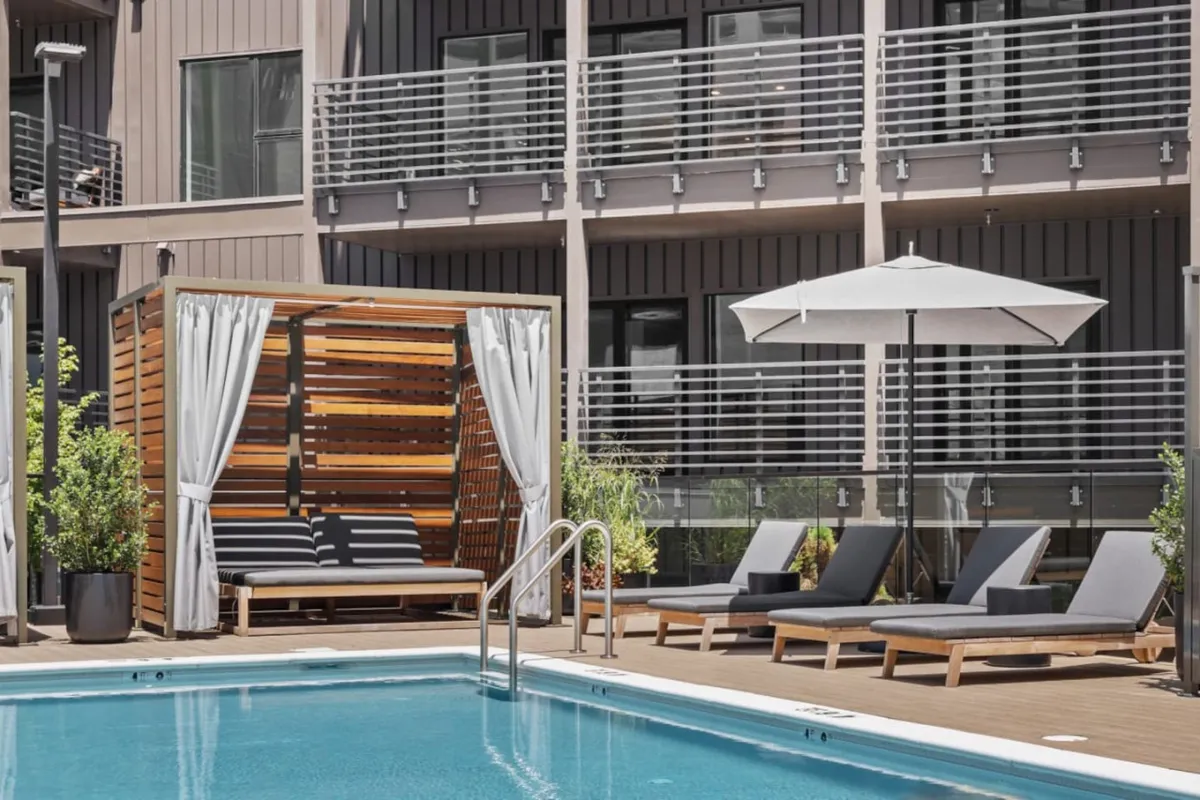 A sparkling swimming pool sits at the heart of the outdoor amenity area, bordered by lounge chairs, shaded cabana seating, and umbrellas for relaxation. The scene is serene yet inviting, designed for both sunbathing and socializing with friends. The cabana area hints at privacy and comfort, while the water glistens under open skies.
