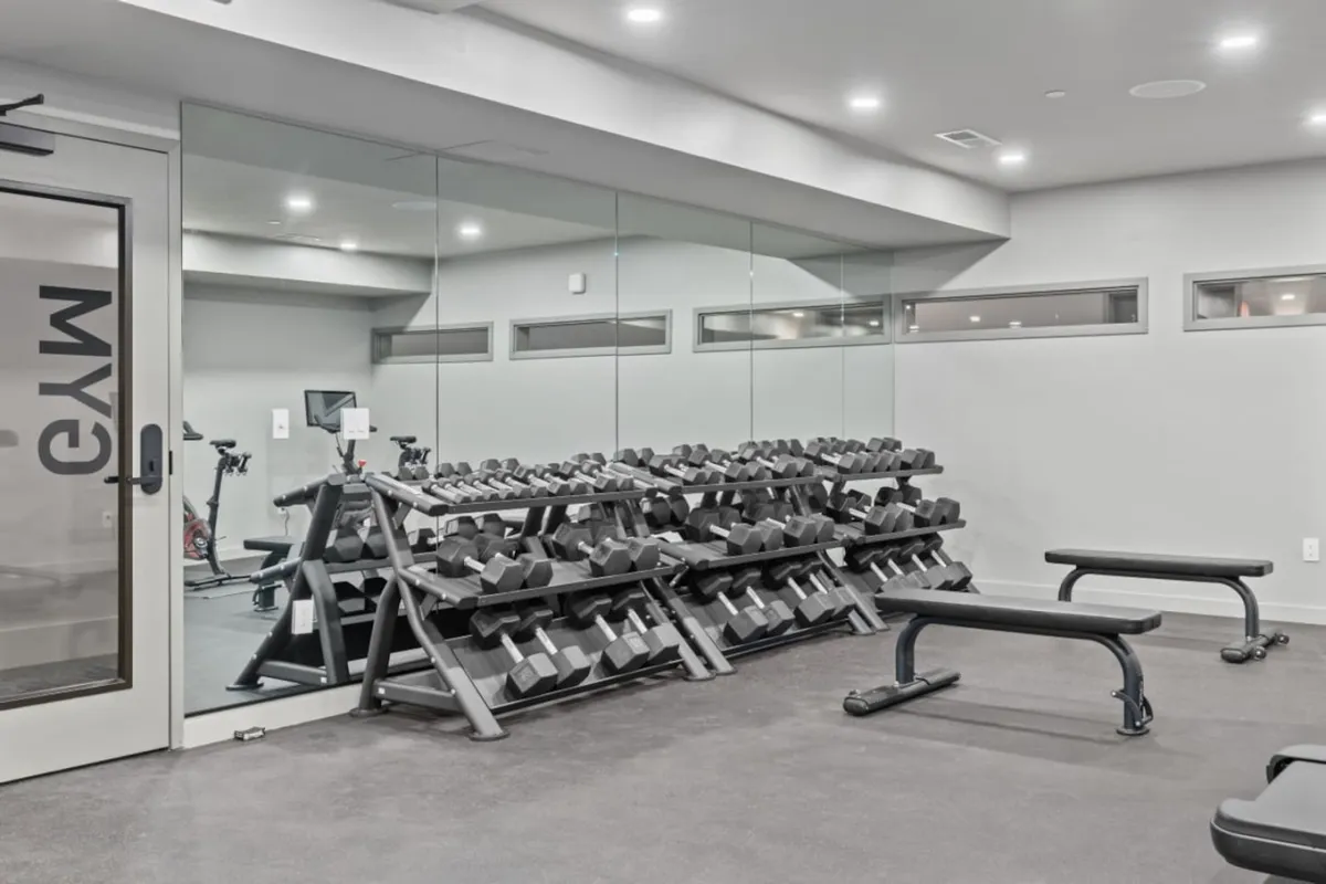 This energetic interior shot highlights a state-of-the-art fitness center equipped with weight racks, benches, and mirrored walls. The space is brightly lit and inspiring, clearly designed to promote health and productivity for residents looking to recharge between meetings or virtual calls.