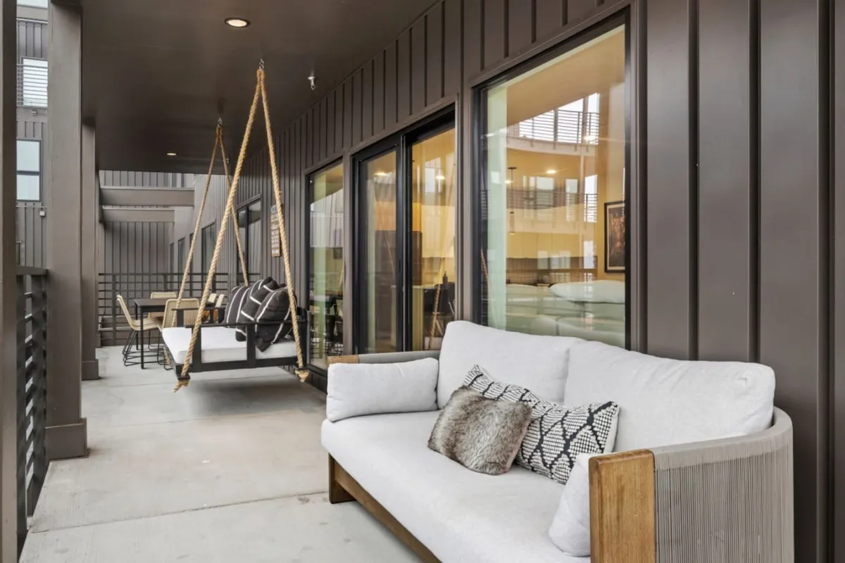 An inviting outdoor lounge area featuring a swinging seat, a modern sofa with textured cushions, and a relaxed, sheltered vibe, perfect for socializing or unwinding. The setting is urban yet green, suggesting a blend of city living and nature.