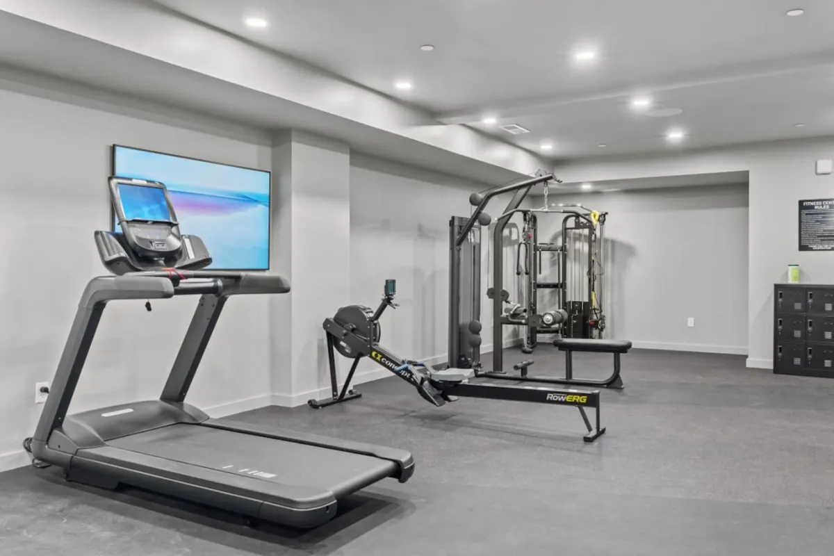 A spacious, well-equipped fitness center featuring a treadmill, rowing machine, weight machine, and a wall-mounted TV, set in a bright and motivational environment. The modern equipment and layout suggest a state-of-the-art gym ideal for cardio, strength training, or guided virtual classes.