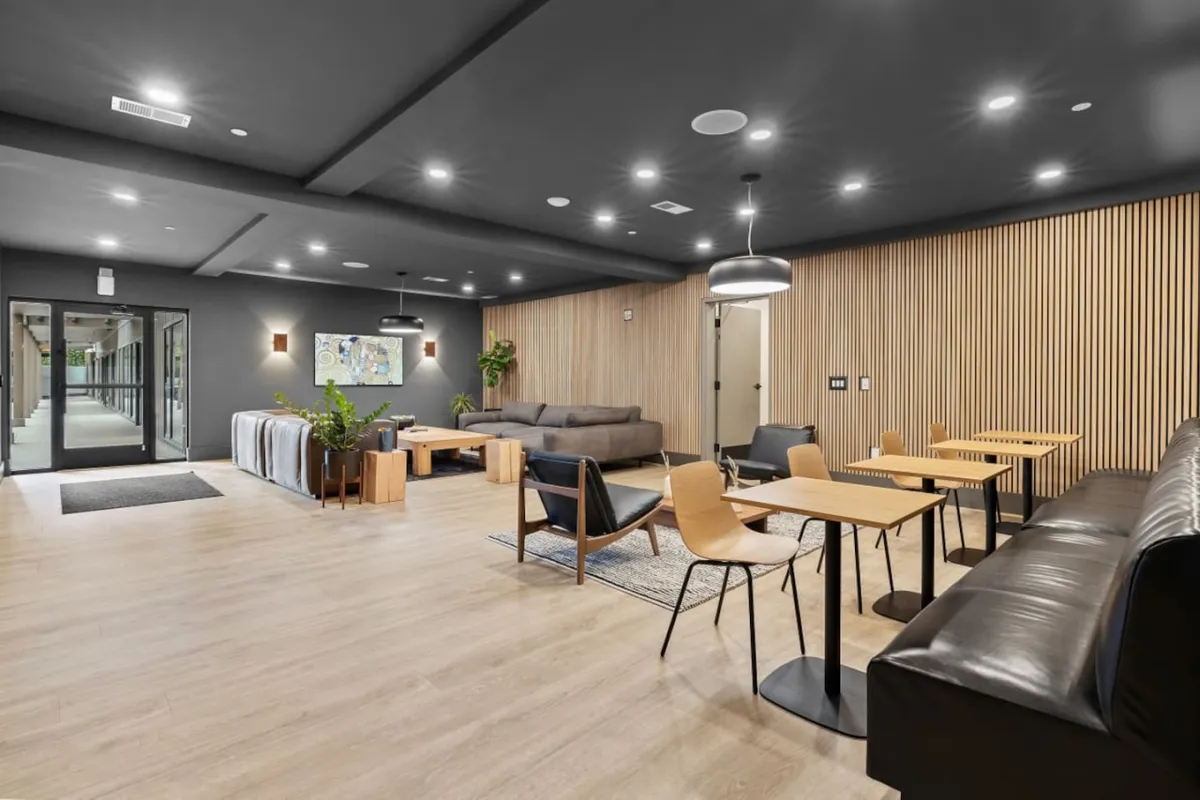 A contemporary community lounge with a comfortable sofa, wooden tables, modern lighting, and plants. The layout invites both productivity and relaxation, with space suitable for casual work, reading, or socializing with colleagues and neighbors.