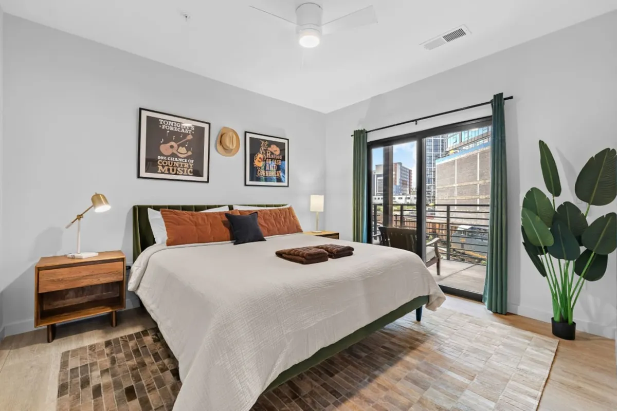A cozy bedroom with green bedding, wall art, an indoor plant, and large windows that open directly onto the balcony. Natural light floods the space, hinting at sunrise views and an easy flow between indoor and outdoor living.
