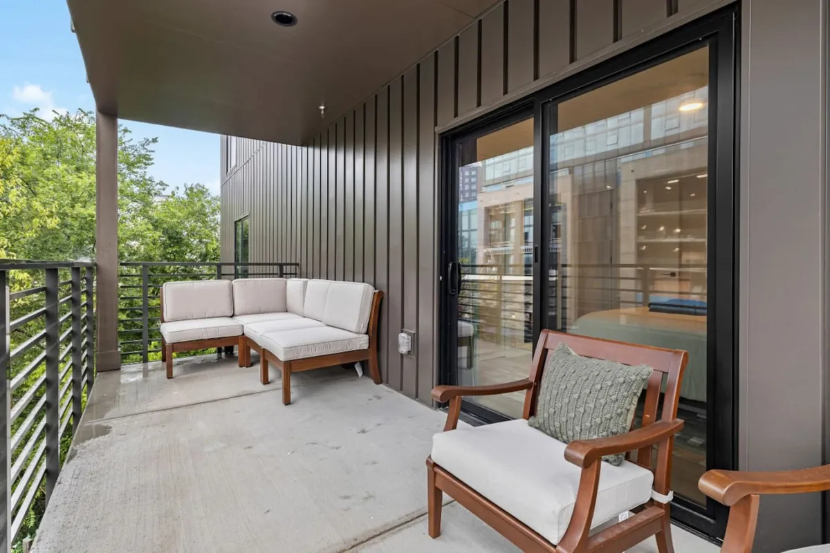 This exterior image features a peaceful outdoor seating area with comfortable chairs and lush greenery. Large glass doors suggest a seamless transition from indoor workspaces to relaxing outdoor environments, perfect for recharging during breaks. The setting is inviting for enjoying a morning coffee or taking a call in the fresh air, reinforcing the blend of work and leisure in a vibrant urban context.