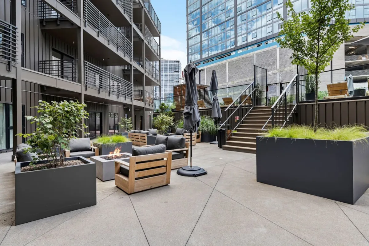 A contemporary outdoor space features a cozy fire pit surrounded by comfortable seating, modern planters, and a staircase. The area is ideal for evening gatherings, relaxation, or reading by the fire as night falls.