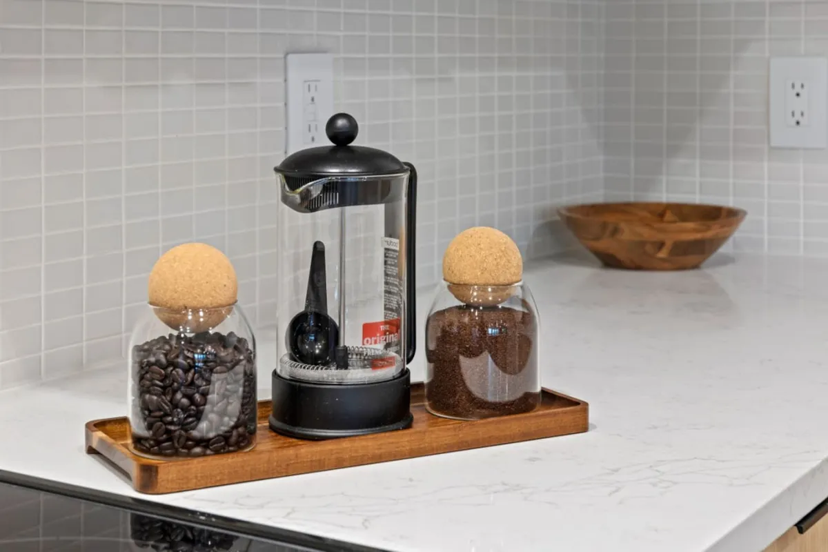 This image focuses on a French press coffee maker, glass jars filled with coffee beans and ground coffee, all beautifully displayed on a wooden tray. The warm lighting and inviting setup suggest a cozy, aromatic environment perfect for morning routines and coffee appreciation.