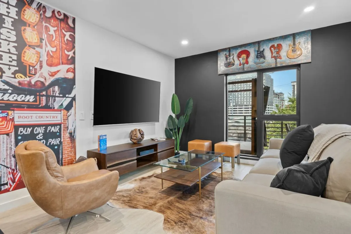 A plush living area featuring a large television, stylish wall decor, a glass coffee table, greenery, and a comfortable sofa. The room exudes a lively yet inviting energy, ideal for gatherings and relaxed evenings with friends.
