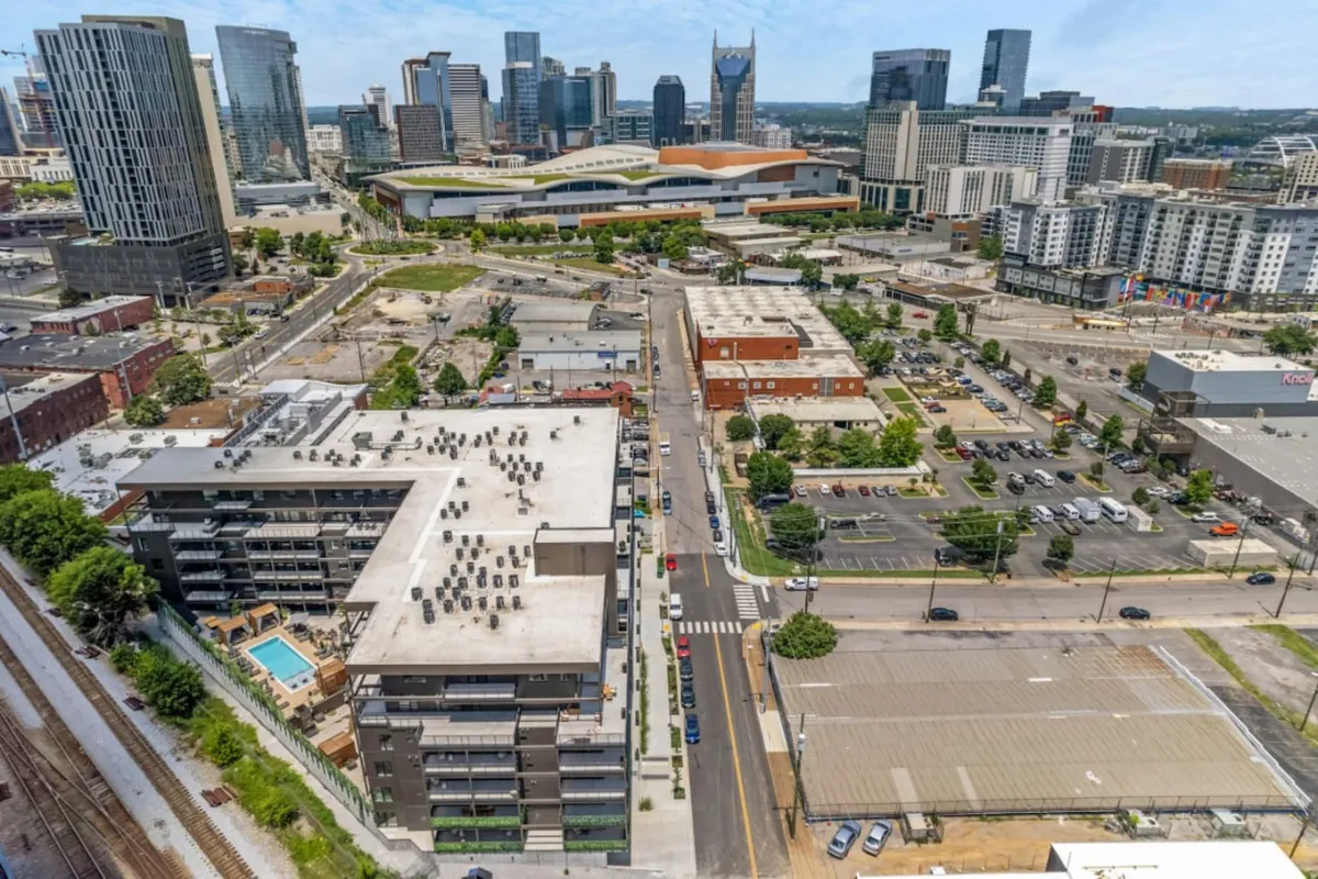 A vibrant rooftop scene featuring a swimming pool surrounded by lounge chairs, with sweeping views of Nashville's modern high-rise skyline. Green spaces and urban infrastructure are visible, hinting at the proximity to major attractions and city life. The setting has an energetic and contemporary vibe, perfect for socializing or relaxing against the city backdrop.