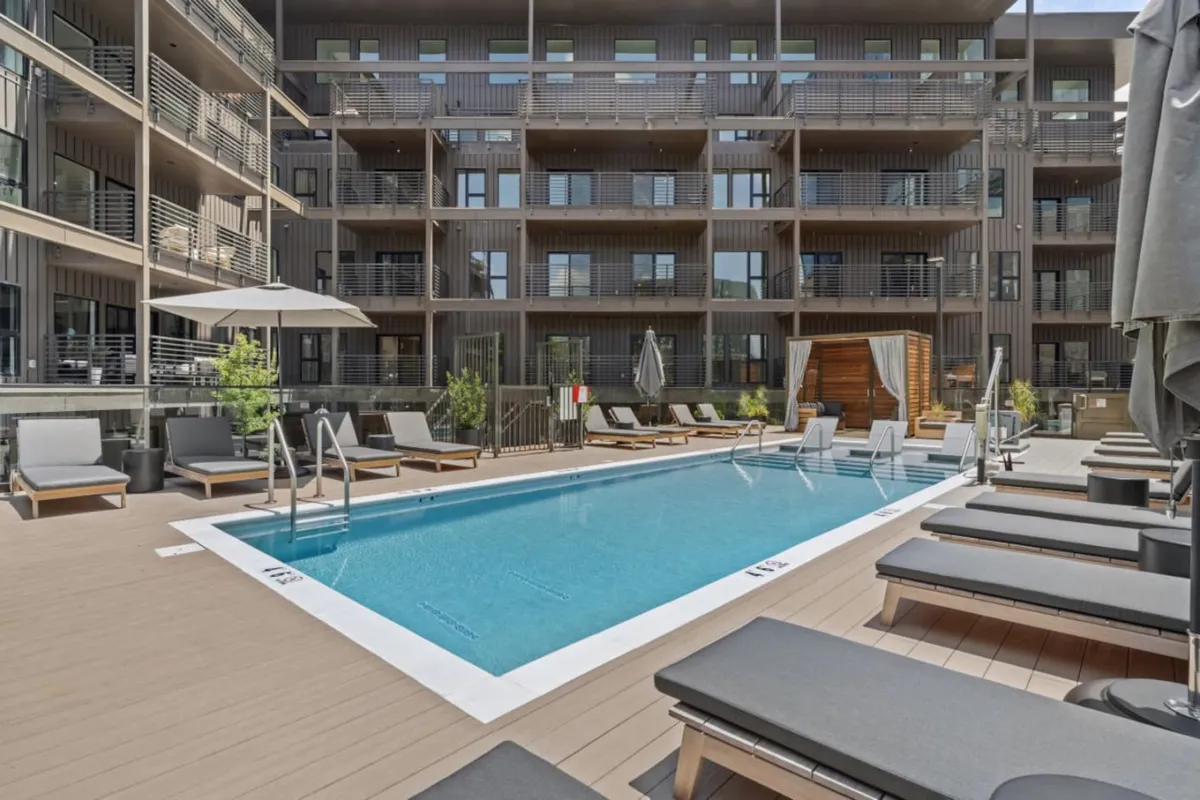 A vibrant outdoor swimming pool with sleek lounge chairs, umbrellas for shade, and lush landscaping. The scene captures a resort-style ambiance, perfect for group relaxation, socializing, or hosting a poolside party, with hints of the city beyond.